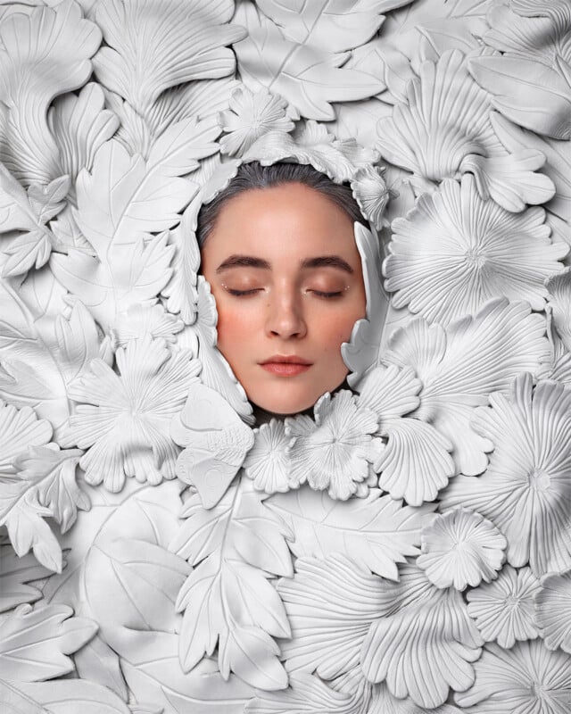 A woman's face with closed eyes emerges from a surrounding of intricately arranged, white, leaf-like sculptural patterns. The monochromatic background highlights her serene expression, blending human features with artistic botanical forms.