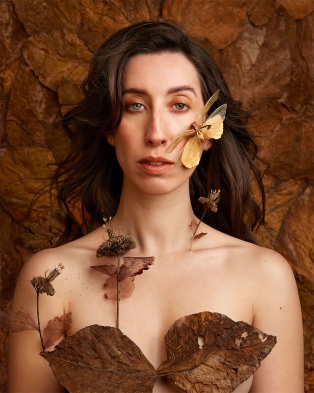 A woman with long dark hair stands against a backdrop of dried leaves. She wears dried leaves and flowers, and a light-colored butterfly rests on her cheek. Her expression is calm, and the earthy tones create a natural, autumnal atmosphere.