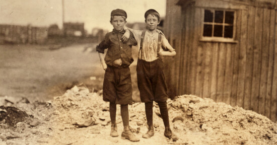 Two young boys stand side by side with their arms around each other. They are dressed in worn, old-fashioned clothing and caps. The background shows a desolate, industrial area with a wooden building on the right and factory structures in the distance.