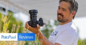 A person with gray-streaked hair and a beard is holding a camera and smiling while looking at it. There is a Petapixel logo and the word "Reviews" superimposed on the bottom left corner. The background shows blurred greenery and outdoor elements.