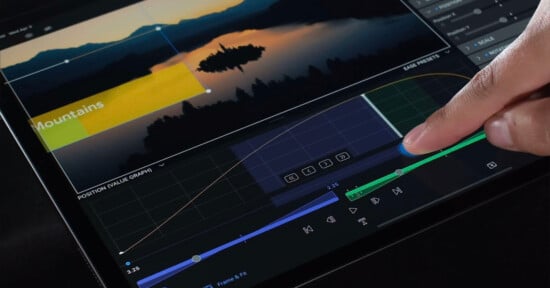 A close-up of a person's finger touching a tablet screen displaying a video editing interface. The screen shows a timeline with various controls, graphs, and a preview of a landscape video featuring mountains and a lake at sunset.