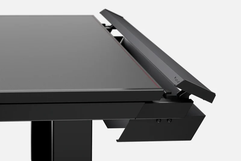 A modern black adjustable desk viewed from an angle, showing its sleek, flat surface and a cable management compartment at the back. The compartment has a lid that can open to organize and conceal cables neatly, contributing to a clean and minimalist design.