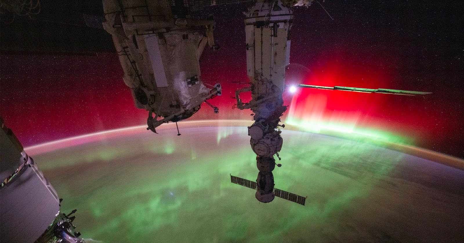 A stunning view from the International Space Station shows a vivid red and green aurora on Earth's horizon with stars in the background. Parts of the space station are visible in the foreground, silhouetted against the colorful lights.