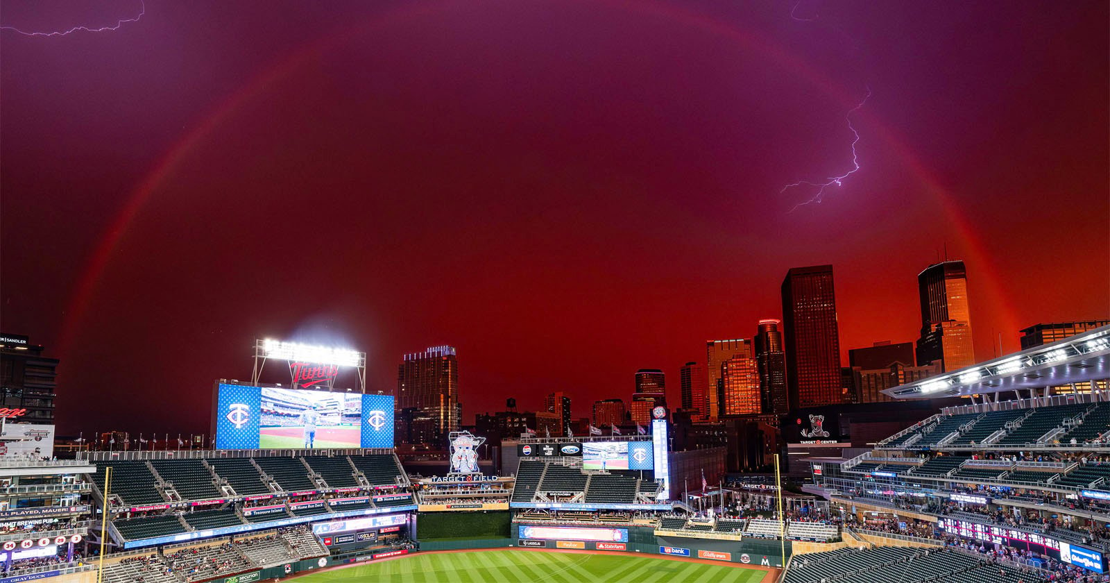 A twilight scene at a baseball stadium shows an impressive rainbow arching across a darkening sky, with a lightning bolt visible near the right side. The city skyline and lit-up stadium scoreboards enhance the dramatic atmosphere.