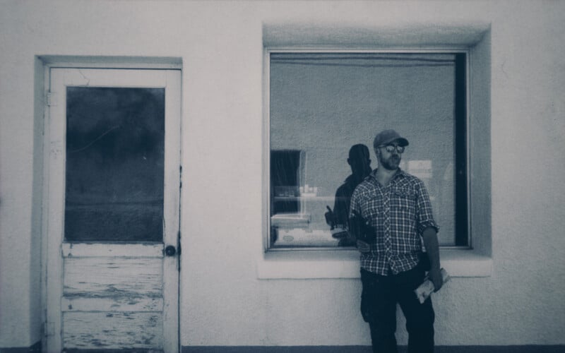 A man in a plaid shirt, baseball cap, and sunglasses leans against a whitewashed wall between a weathered door and a large window. The photo is black and white, and he holds an object in his left hand while looking off to the side.