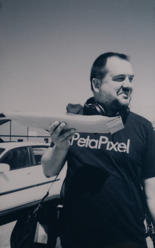 A person stands outdoors holding a paper airplane. They are wearing a "PetaPixel" T-shirt and a pair of headphones around their neck. A car is visible in the background, parked next to a building. The image is in black and white.