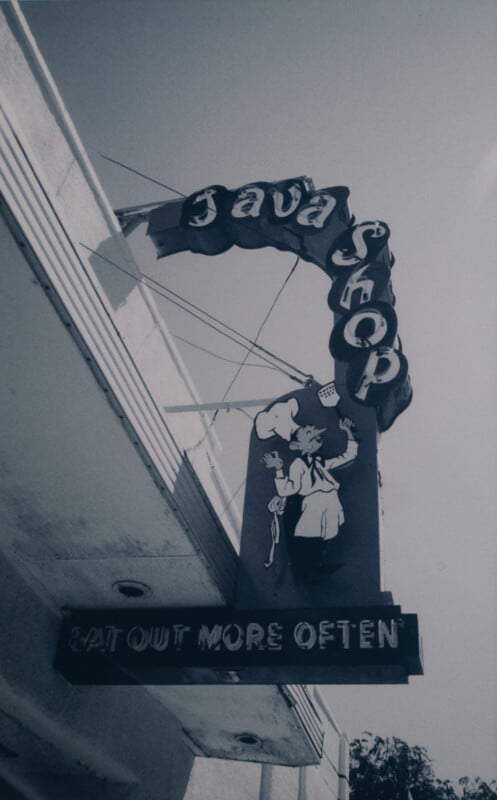 A vintage sign for a Java Shop hangs from the side of a building. The sign includes an illustration of a chef holding a steaming cup of coffee. Below the main sign, another sign reads, "EAT OUT MORE OFTEN." The image is in black and white.