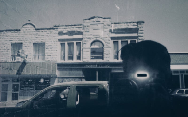 A grainy black-and-white photograph depicting the reflection of a person holding a camera in a store window. In the background, there is an old stone building with arched windows and a truck parked in front of it.