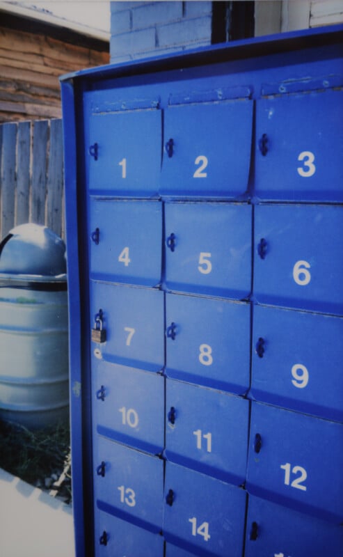 A blue metallic mailbox unit with numbered compartments ranging from 1 to 14, positioned against a wooden fence and adjacent to a gray trash bin. The top of the unit is slightly rusted and the mailbox appears to be outdoors in daylight.