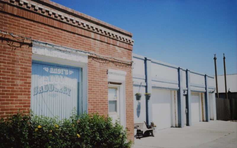 A brick building with a sign that reads "Andy’s Saddle & Tack" on the window. Next to it, a light blue building with three garage doors. A small bush with yellow flowers and a black bench are in front of the buildings. The sky is clear and blue.