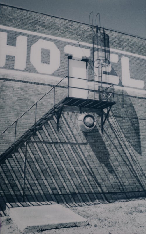 A black-and-white photograph of a brick building with the word "HOTEL" painted on it. A metal staircase leads to a door midway up the wall, with shadows cast by the staircase creating a diagonal pattern on the brick surface. A metal ladder extends above the door.