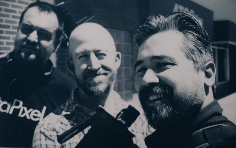 Three men are smiling at the camera in an outdoor setting with a brick wall and buildings in the background. The man in the foreground is taking a selfie, holding the camera stick. It's a sunny day with clear visibility.