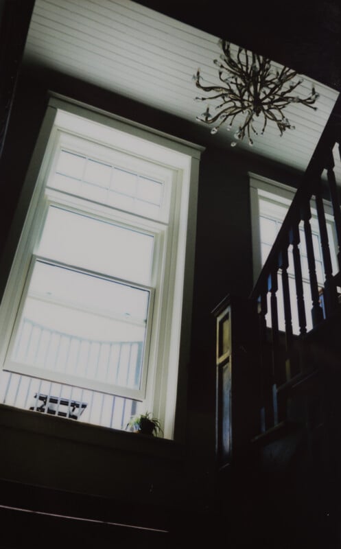 A softly lit staircase ascends to a landing with two large, vertically aligned windows illuminating the area. A chandelier with intricate designs hangs from the ceiling. Sunlight streams through the windows, casting a glow on the wooden stairs and railing.