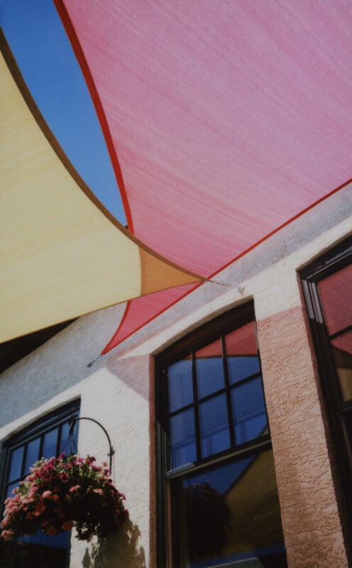 A brightly colored fabric canopy, featuring pink and yellow shades, stretches overhead against a clear blue sky. Below, a white stucco building with large windows and a hanging basket filled with pink flowers can be seen.