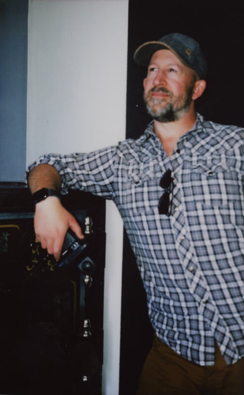 A man with a beard wearing a checkered shirt and a baseball cap leans against a dark-colored wall. He has a smartwatch on his left wrist, and sunglasses attached to his shirt. He's holding a drink can in his left hand and appears to be smiling slightly.