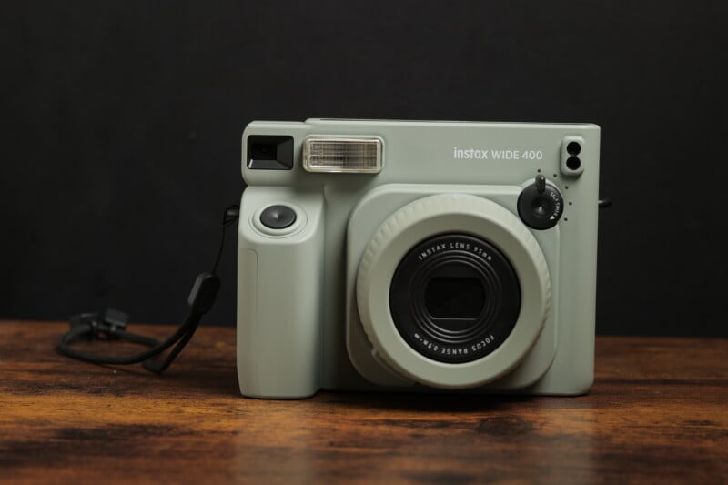 A gray Instax Wide 400 instant camera sits on a wooden surface against a dark background. The camera features a large lens, built-in flash, and various control buttons.