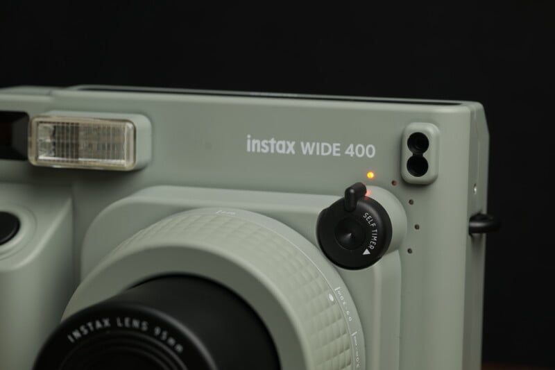 Close-up of an Instax WIDE 400 instant camera in a grayish-green color. The camera features an automatic flash, adjustable lens, shutter button, and various controls. The ON/OFF switch is indicated by a small orange light on the front.