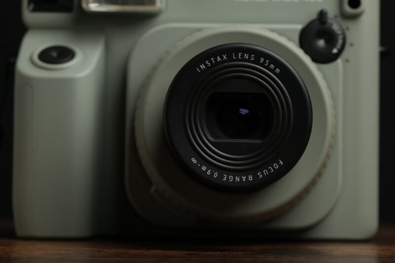 A close-up photo of a light green instant camera, focusing on the lens. The lens is labeled "INSTAX LENS 95mm" with "FOCUS RANGE 0.9m" written on it. The body of the camera features buttons and a flash near the lens.