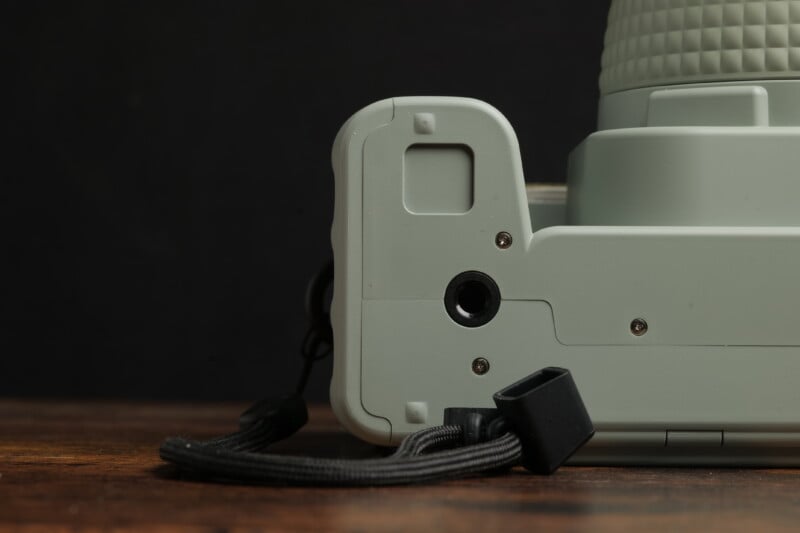 Close-up view of the bottom-left corner of a light green camera resting on a dark wooden surface. The image showcases part of the camera's body, a small lanyard attached to it, and a black background.