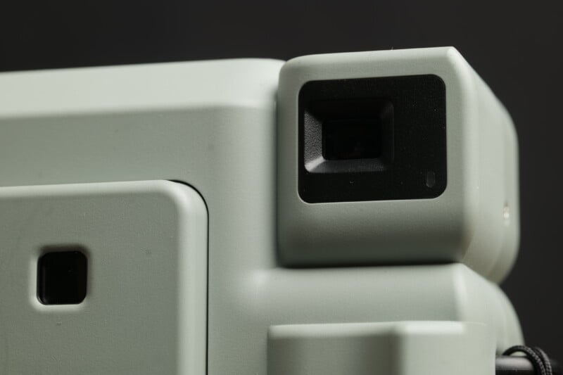 A close-up photo of the viewfinder and body of a retro-style instant camera. The camera features a textured, light green exterior with a small black viewfinder and a slightly protruding flash unit on the right side. The background is blurred and dark.