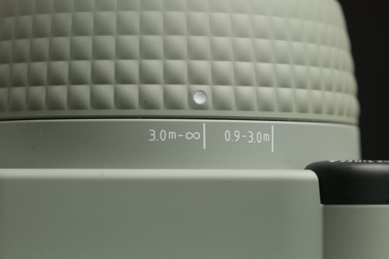 Close-up image of a camera lens displaying focus distance markings. The markings indicate two focus ranges: "3.0m to infinity" and "0.9 to 3.0m." The lens surface has a textured, grid-like pattern.