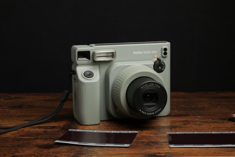 A light gray Instax Wide 400 instant camera is placed on a wooden surface against a dark background. The camera has a strap attached to it and two undeveloped instant film photos are laid in front of it.