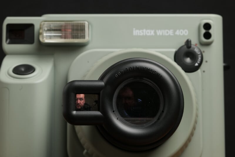 Close-up of a light gray Instax WIDE 400 camera with a reflective lens. A small part of a person's face is visible in the reflection of the camera lens, showing their eyes and forehead. The background is black.