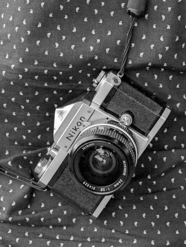 Black and white photo of a vintage Nikon film camera with a strap, placed on a patterned fabric background. The camera has a manual lens and is positioned at a slight angle.