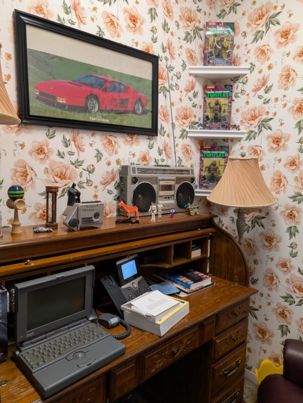 A vintage home office with floral wallpaper features a wooden desk with an old laptop, phone, papers, and small figurines. A framed car print hangs above a retro boombox. Shelves display Teenage Mutant Ninja Turtles action figures. A lamp sits on the desk.