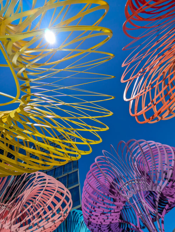 A view from below of an art installation featuring large, colorful, spiral sculptures set against a clear blue sky. The sculptures are yellow, orange, purple, pink, and blue, and create an intricate pattern as they intertwine. The sun shines brightly in the background.