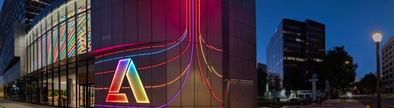 Wide-angle view of an urban street at night with modern buildings. The building on the left features colorful illuminated lines and a prominent, multi-colored "A" logo on its facade. Streetlights and trees line the road on the right side.