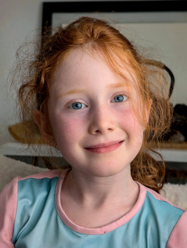 A young girl with curly red hair and blue eyes is smiling softly at the camera. She is wearing a blue and pink shirt. The background appears to be indoors, with a blurred dark frame and some objects out of focus.