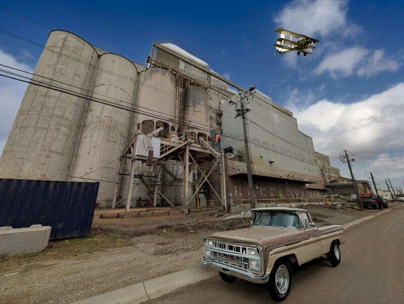 A vintage pickup truck is parked in front of a large, industrial concrete building with silos. Overhead, a yellow biplane is flying in a partly cloudy blue sky. Power lines and a fence are visible in the foreground.