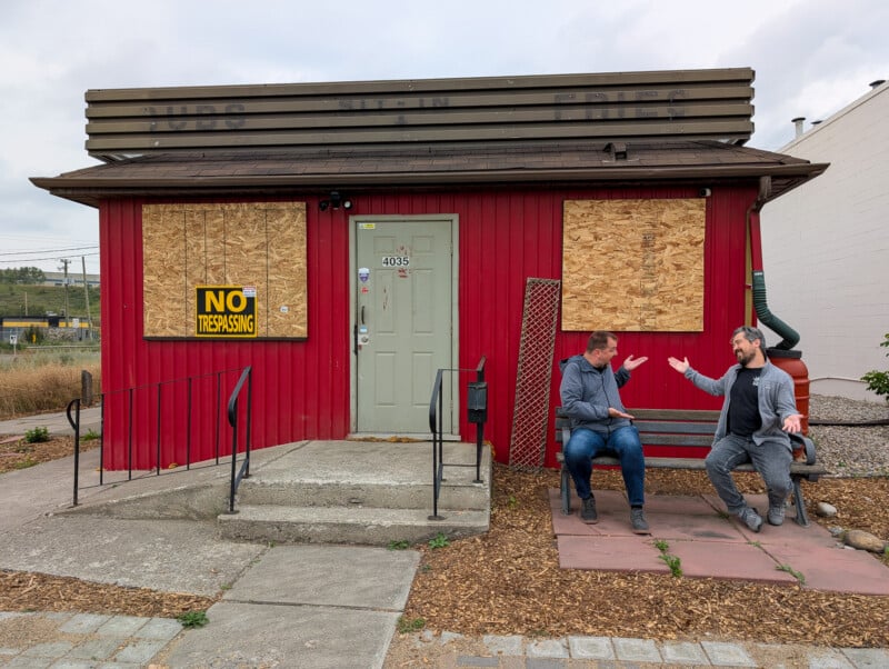 Two men are engaged in a lively conversation in front of a small, boarded-up red building with a "No Trespassing" sign. One man sits on a bench, and the other stands with animated gestures. The scene appears casual and informal.