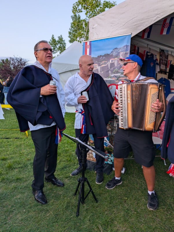 Three men dressed in traditional attire interact at an outdoor event near a tent with cultural items displayed. Two hold drinks while one plays an accordion. The surroundings include grass, trees, and additional decorations, evoking a festive atmosphere.