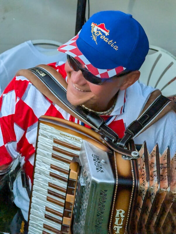 A man wearing a Croatia cap and sunglasses plays an accordion. He is smiling and dressed in a red and white checkered shirt. The accordion has intricate designs on its body. The background is slightly blurred, showing part of a white chair.