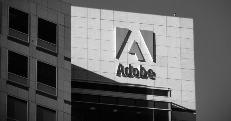 Black and white photo of a modern building with the Adobe logo prominently displayed on its facade. The building has large, square windows and the sleek design typical of corporate architecture. The Adobe logo consists of a stylized 'A' and the word "Adobe".