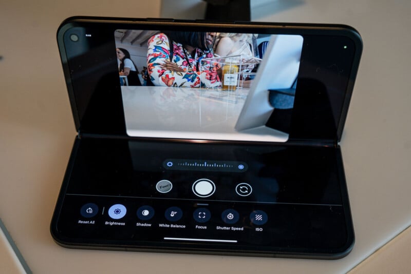 A foldable smartphone is partially open and displaying a camera interface. The top screen shows a photo being taken of a person in colorful clothing at a table with drinks. The bottom screen displays various camera controls like brightness, focus, and shutter speed.