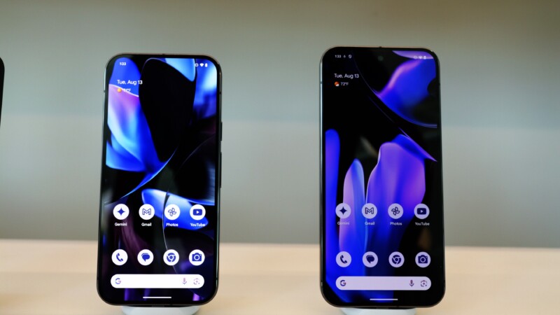 Two smartphones are on display side by side against a light background. Both screens are lit up, showing an abstract blue and black patterned wallpaper. The home screens feature various app icons, including camera, Gmail, Photos, and YouTube.