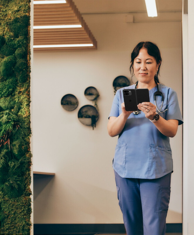 A healthcare professional in blue scrubs and a stethoscope around her neck is standing indoors and looking at a smartphone. There is a green plant wall on the left and some decorative wall plates in the background.