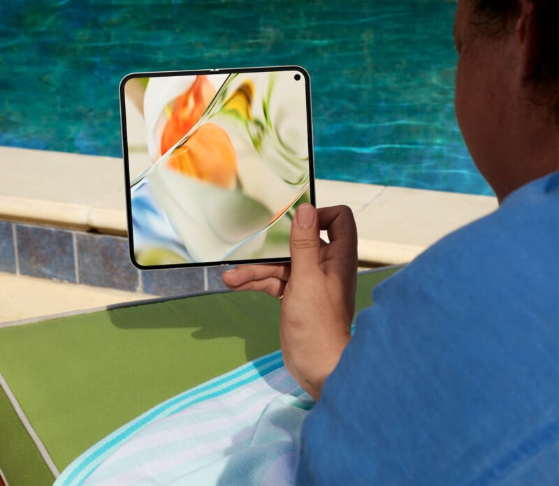 A person wearing a blue shirt and holding a tablet with a vibrant abstract design on the screen while sitting on a green lounge chair. The scene is set by a pool with clear blue water in the background.