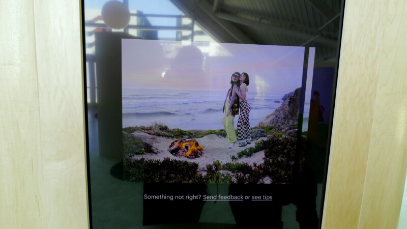 A photograph displayed on a screen shows two individuals standing together in front of a scenic beach with waves and cliffs in the background. There is a small campfire near them. The screen has a message at the bottom: "Something not right? Send feedback or see tips.