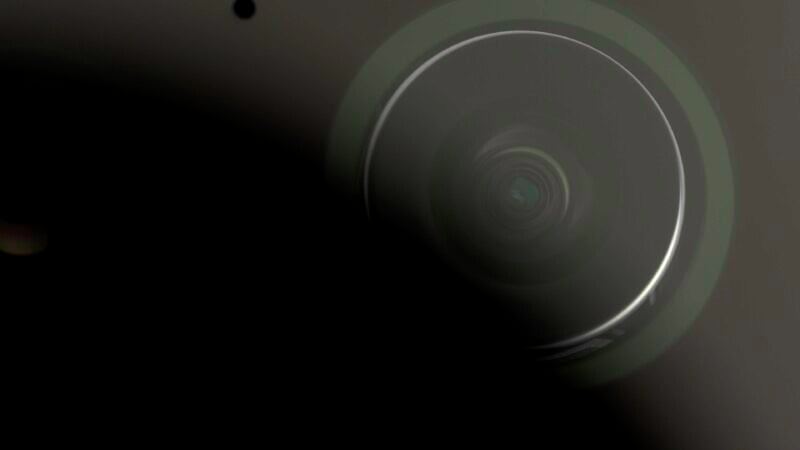 Close-up image of a camera lens with a circular, glowing ring around it. The background is mostly dark and shadowy, with parts of the lens and camera body faintly visible.