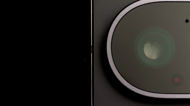 Close-up view of a camera lens on a dark background. The lens is large and round, surrounded by a circular frame. The image highlights the minimalistic and sleek design of the camera.