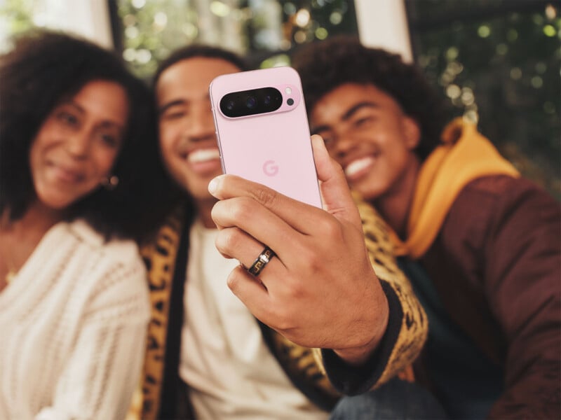 Three people are smiling as they take a selfie with a pink smartphone. The phone has a "G" logo on the back. They appear to be indoors, with a window and greenery visible in the background.