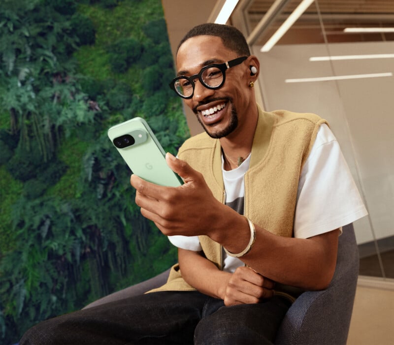 A man wearing glasses, a beige vest over a white shirt, and wireless earbuds, is sitting on a chair indoors. He is smiling broadly while looking at his green smartphone. In the background, there is a wall covered with green foliage.