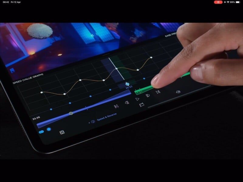 Close-up of a person interacting with a tablet's screen, which displays a video editing software with various graphs, timelines, and controls. The person's finger is tapping a green play button on the interface. The background shows a colorful, out-of-focus scene.