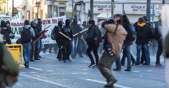Protesters in a street demonstration hold banners and wooden sticks. Many participants wear masks and dark clothing. The scene is tense, with some individuals appearing to clash. In the background, shops and signs are visible.