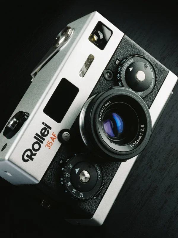 A close-up image of a vintage Rollei 35AF camera. The camera features a black and silver body, with a prominently displayed lens labeled "RolleiLens f=38mm 1:2.8". Various dials and buttons for settings and adjustments are visible on the camera's body.