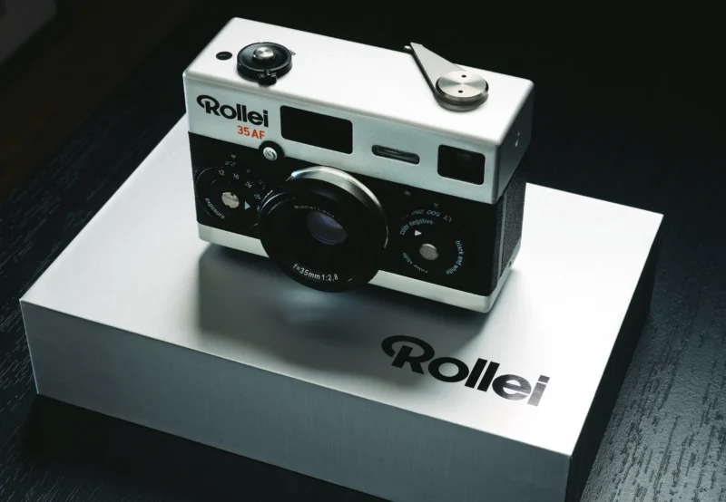 A Rollei 35 AF film camera is displayed on a matching Rollei-branded box. The camera features a classic design with a silver and black finish, manual control dials, and a lens at the center. The background is a reflective dark surface.